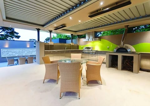 Modern outdoor kitchen with green backsplash, stainless steel appliances, woven chairs, and covered patio with ambient lighting