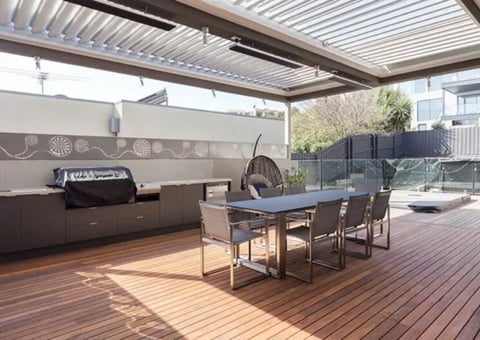 Modern outdoor patio with pergola roof, built-in grill, dining table, chairs, and wooden deck flooring under bright sunlight